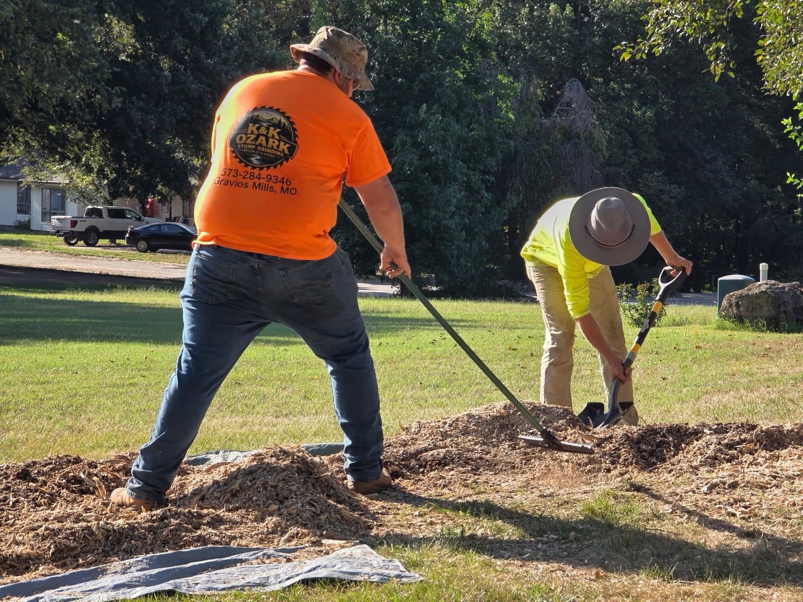 Two K&K Ozark crew members in orange shirts raking and shoveling fresh stump grindings on a lawn